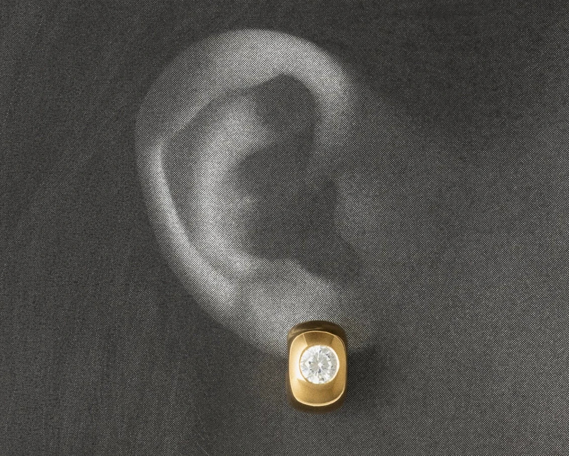 A close-up of a human ear wearing a small gold stud earring with a clear gemstone, set against a dark textured background.