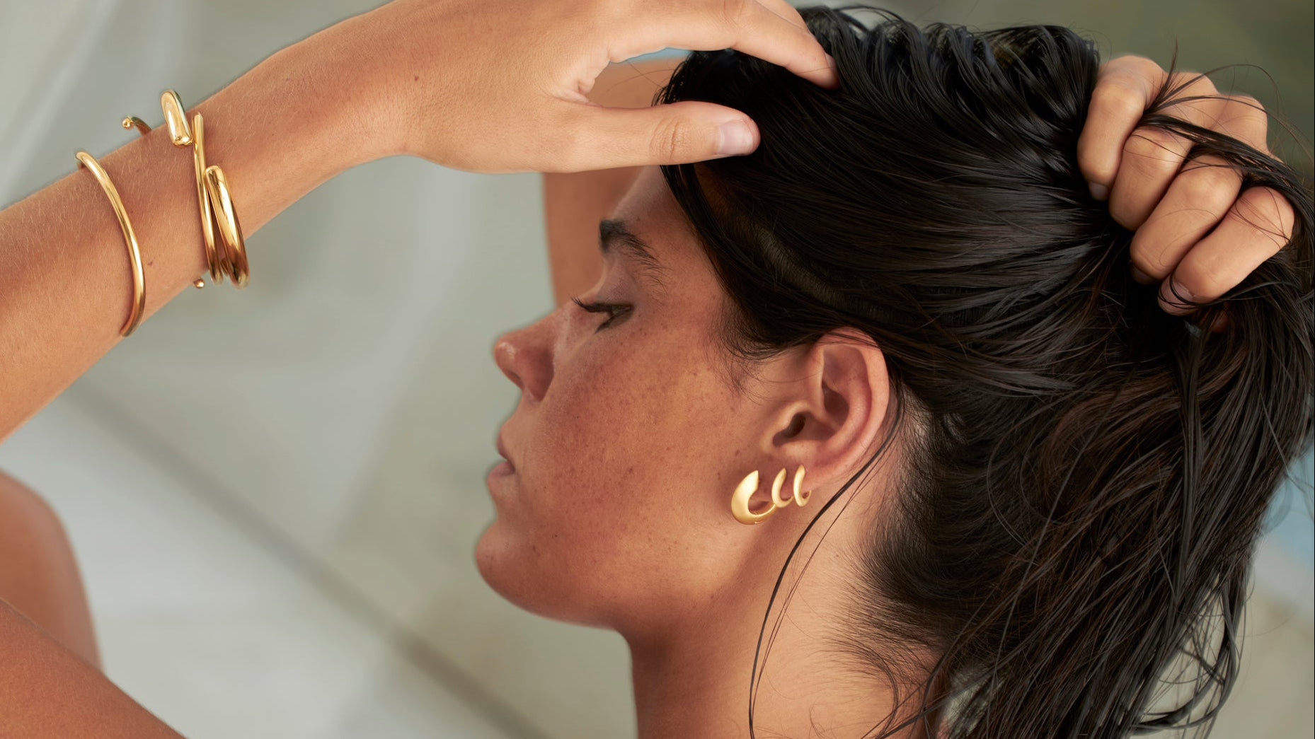Woman with gold earrings and bracelets adjusting her hair against a neutral background
